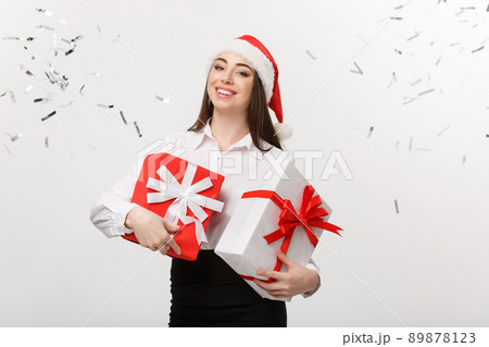 Business Concept - Beautiful young caucasian business woman with santa hat holding gift box with confetti celebration background. Business Concept - Beautiful young caucasian business woman with santa hat holding gift box with confetti celebration background. 89878123