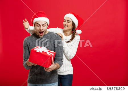 Christmas Concept - Young woman covering man's eyes with hand and giving surprise big gift. Isolated on Red background Christmas Concept - Young woman covering man's eyes with hand and giving surprise big gift. Isolated on Red background 89878809