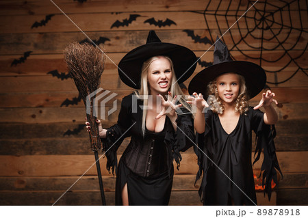 Halloween Concept - cheerful mother and her daughter in witch costumes celebrating Halloween posing with curved pumpkins over bats and spider web on Wooden studio background. Halloween Concept - cheerful mother and her daughter in witch costumes celebrating Halloween posing with curved pumpkins over bats and spider web on Wooden studio background. 89878918