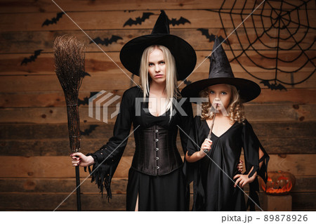 Halloween Concept - cheerful mother and her daughter in witch costumes celebrating Halloween posing with curved pumpkins over bats and spider web on Wooden studio background. Halloween Concept - cheerful mother and her daughter in witch costumes celebrating Halloween posing with curved pumpkins over bats and spider web on Wooden studio background. 89878926