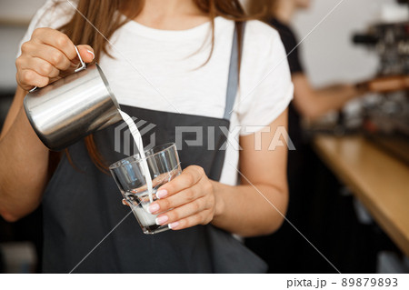 Coffee Business Concept - close-up lady barista in apron preparing and pouring milk in glass cup while standing at cafe. Coffee Business Concept - close-up lady barista in apron preparing and pouring milk in glass cup while standing at cafe. 89879893