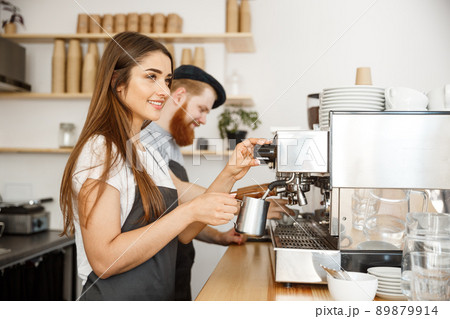 Coffee Business Concept - portrait of lady barista in apron preparing and steaming milk for coffee order with her partner while standing at cafe. Coffee Business Concept - portrait of lady barista in apron preparing and steaming milk for coffee order with her partner while standing at cafe. 89879914
