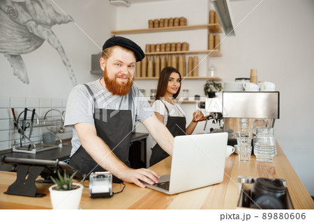 Coffee Business Concept - Young handsome bearded bartender, barista or manager working and planing in laptop at modern coffee shop. 89880606