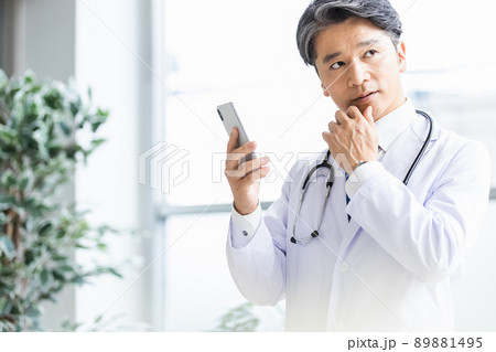 スマホを持って窓際に立つ白衣の医師 89881495
