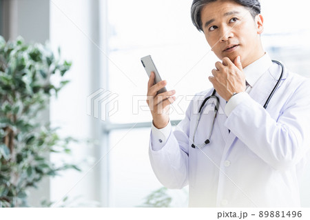 スマホを持って窓際に立つ白衣の医師 89881496