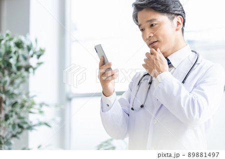 スマホを持って窓際に立つ白衣の医師 89881497