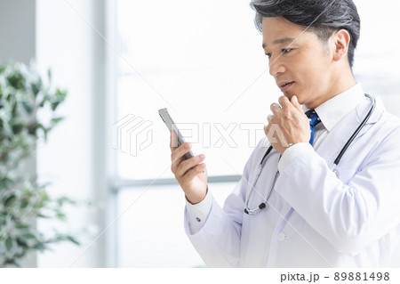 スマホを持って窓際に立つ白衣の医師 89881498