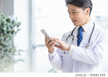 スマホを持って窓際に立つ白衣の医師 89881499