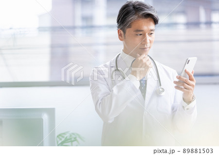 スマホを持って窓際に立つ白衣の医師 89881503