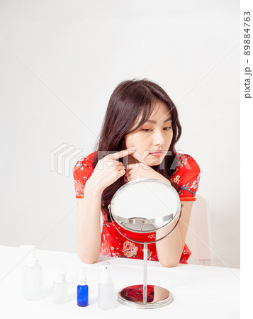 Portrait of a young asian woman in traditional cheongsam qipao dress looks at mirror and does skin care isolated on white background. 89884763