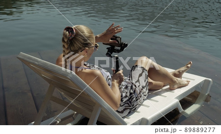 Woman shooting on handheld film gimbal stabilization for smartphone. Girl lie sunbed on pier make selfie. Blogger broadcast video vlogging. 89889277