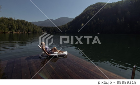 Woman lie on a sunbed in sunglasses and a boho silk shawl. Girl rest on a flood wood underwater pier. The pavement is covered with water in the lake. 89889318