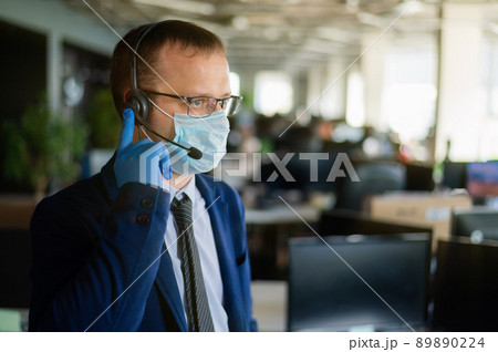 A man in latex gloves and a medical mask is talking to a client over the headset. A male office manager answers calls. Work during the coronavirus. White-collar tie and jacket. A man in latex gloves and a medical mask is talking to a client over the headset. A male office manager answers calls. Work during the coronavirus. White-collar tie and jacket. 89890224