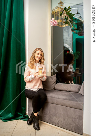 Beautiful caucasian young smiling woman with long wavy blonde hair holding and drinking a latte from a glass cup, sitting on a sofa 89892987
