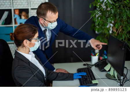 Colleagues in surgical masks in an open office space communicate at the work desk. A male top manager teaches a female intern how to work at a computer. The boss directs the subordinate. 89893123
