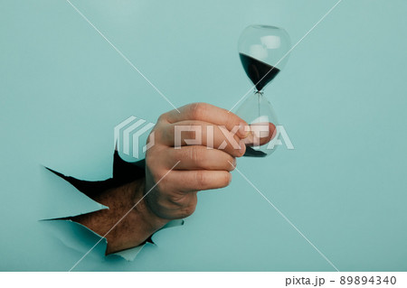 Male hand holds hpurglass in torn hole of blue background Male hand holds hpurglass in torn hole of blue background 89894340