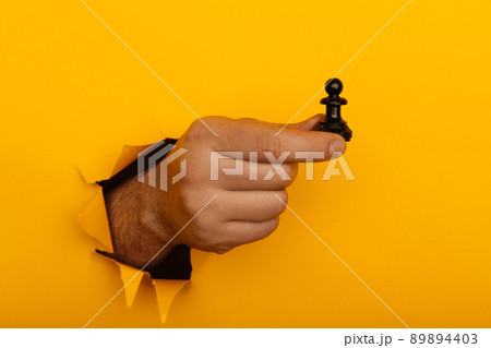 Hand is holding figure of pawn through a hole of yellow background 89894403