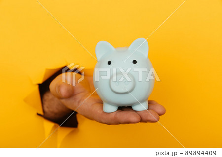Savings concept. Hand with a blue pink piggy bank through a hole in yellow background close-up 89894409