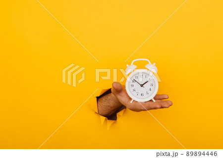 Hand holds alarm clock through a paper hole in yellow background with copy space Hand holds alarm clock through a paper hole in yellow background with copy space 89894446