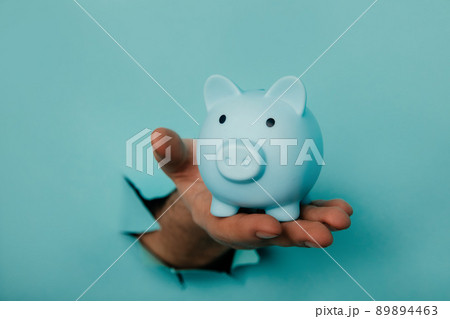 Hand with a blue piggy bank through a blue paper hole. Financial and business concept Hand with a blue piggy bank through a blue paper hole. Financial and business concept 89894463