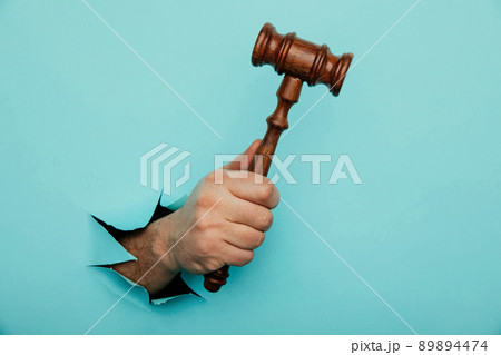 Hand holding a judge's gavel through torn blue paper wall. Law and courts, triumph of justice 89894474