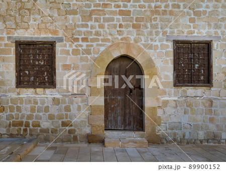 Old weathered arched wooden door and two closed rusted wrought iron windows on bricks stone wall 89900152
