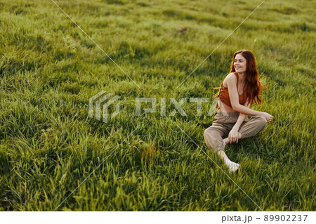 The sunset light of a summer evening illuminates a beautiful young redheaded woman sitting on the grass in green pants and an orange top and smiling at the sun 89902237