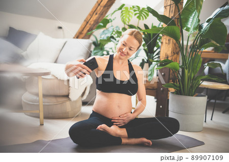 Young happy and cheerful beautiful pregnant woman taking selfie with her mobile phone while staying fit, sporty and active on her maternity leave. Motherhood, pregnancy, yoga concept. 89907109