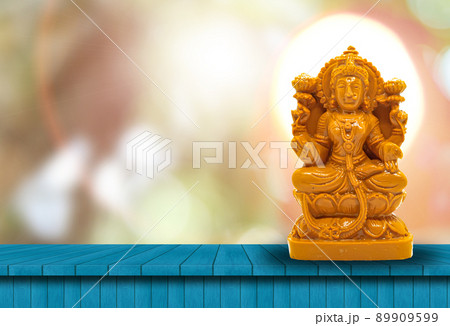 The statue of Mahalakshmi carved in brown wood on a wooden table has a natural bokeh background with a powerful light behind the head 89909599