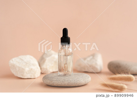 Transparent glass cosmetic dropper standing on stone with dry flowers on beige background. Beauty composition. Concept of natural and original skincare products presentation.  89909946