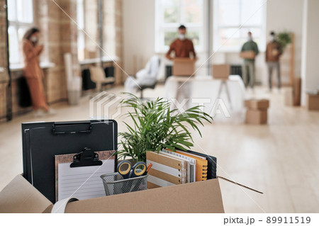 Focus on open cardboard box with office supplies, clipboards and plant against people moving into new office in background 89911519