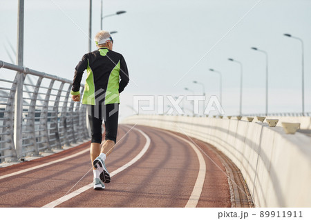 Horizontal back view shot of senior man wearing black and green tracksuit running along track 89911911