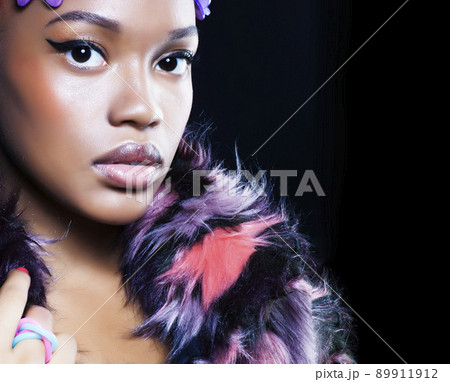 young pretty african american woman in spotted fur coat and flowers on head smiling on black background, fashion people concept close up 89911912