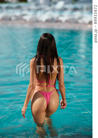 Graceful long haired woman entering in swimming pool at hot sunny day. Back view of slim attractive brunette lady in pink bikini getting into clear turquoise water of luxury resort. Concept of relax. 89933872