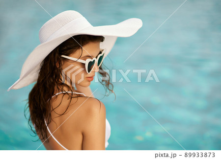 View from shoulder of romantic girl in white hat sunbathing on vacation outdoors. Nice female tourist with wet hair wearing sunglasses with heart shapes relaxing at midday. Concept of vacation. 89933873