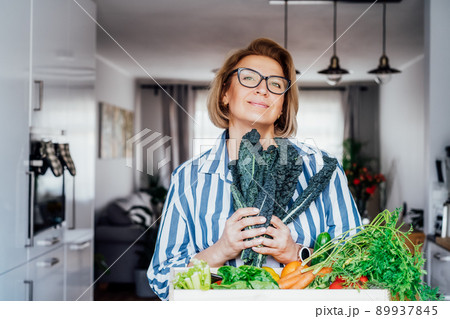 Smiling woman holding fresh kale from her healthy food delivery. Wooden box with vegetables. Support Local farmer food. Start of a healthy life, weight loss concept. Online home food delivery. 89937845