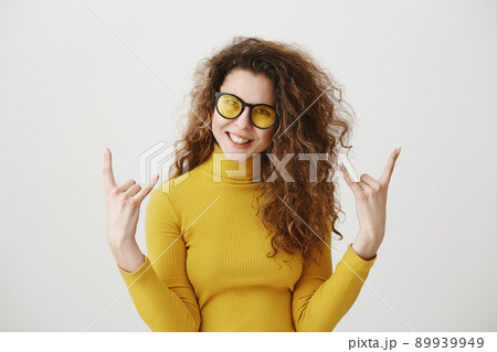 Beautiful funny amazed rocker girl with curly hairstyle and standing with rock sign and looking at camera. studio shot, isolated on grey background 89939949