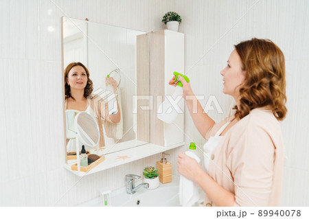 an attractive housewife washes the bathroom mirror with a special glass cleaner. an attractive housewife washes the bathroom mirror with a special glass cleaner. 89940078