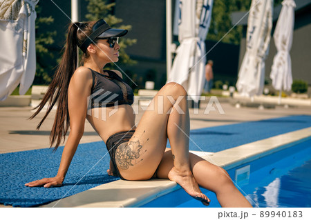 Portrait of a female having rest and posing in a swimming pool. Dressed in a black swimsuit, sunvisor cap and sunglasses. Portrait of a female having rest and posing in a swimming pool. Dressed in a black swimsuit, sunvisor cap and sunglasses. 89940183