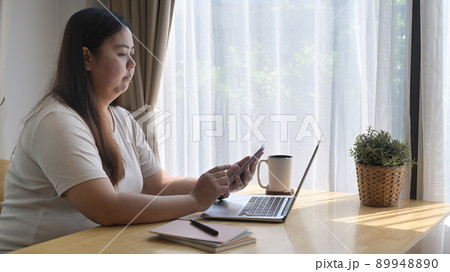 Side view chubby asian woman sitting in front of laptop computer and using mobile phone at home 89948890