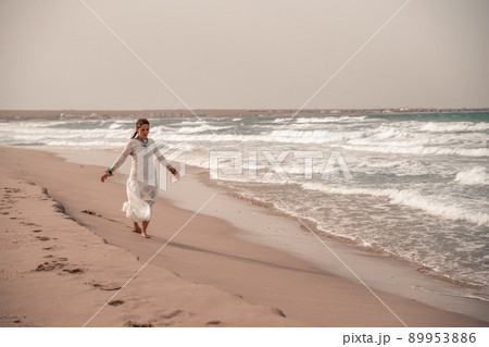 Model in boho style in a white long dress and silver jewelry on the beach. Her hair is braided, and there are many bracelets on her arms. Model in boho style in a white long dress and silver jewelry on the beach. Her hair is braided, and there are many bracelets on her arms. 89953886