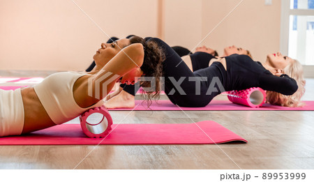 A group athletic women doing pilates or yoga on pink mats in front of a window in a beige loft studio interior. Teamwork, good mood and healthy lifestyle concept. 89953999