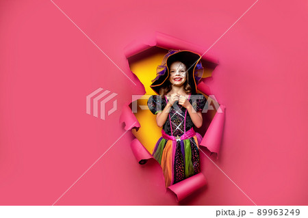 Laughing funny child girl in a witch costume of halloween looking, smiling and scares through a hole of red, yellow paper background. Copyspace Laughing funny child girl in a witch costume of halloween looking, smiling and scares through a hole of red, yellow paper background. Copyspace 89963249