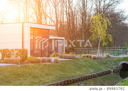 Modern country house with the large lawn and a wooden fence. In front of the house there is a covered terrace with a lounge zone. On the lawn there is a trees and flowerbeds. It is sunny. Outside. Modern country house with the large lawn and a wooden fence. In front of the house there is a covered terrace with a lounge zone. On the lawn there is a trees and flowerbeds. It is sunny. Outside. 89963762