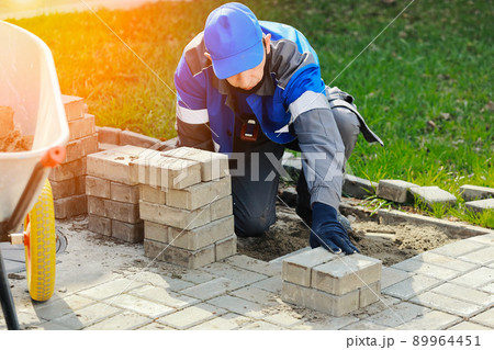 Bricklayer in work clothes sits on sidewalk and lays out paving slabs. Sight of working man in open 89964451