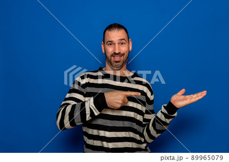 Surprised man in striped sweater pointing left with his finger, shouting WOW, isolated on blue background with copy space for your text. shock content. 89965079