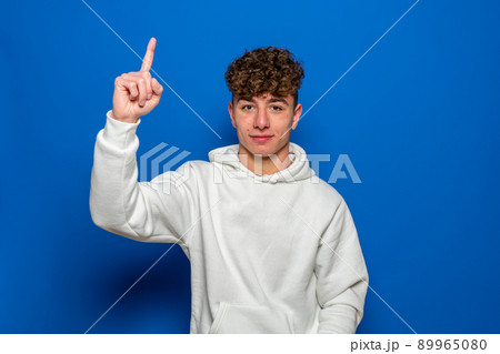 Caucasian young man with curly hair dressed in a white sweatshirt pointing the index finger up, isolated on blue studio background. Caucasian young man with curly hair dressed in a white sweatshirt pointing the index finger up, isolated on blue studio background. 89965080