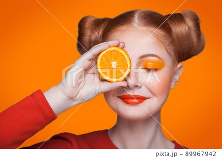 Young beautiful funny fashion model with orange slice on orange background. with orange makeup and hairstyle and freckles. holding orange between eyes with nice smile. Young beautiful funny fashion model with orange slice on orange background. with orange makeup and hairstyle and freckles. holding orange between eyes with nice smile. 89967942