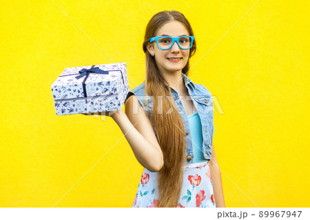 Portrait of a happy hipster smiling girl in dress and blue glasses, holding present box and toothy smile isolated over yellow background. Indoor studio shoot Portrait of a happy hipster smiling girl in dress and blue glasses, holding present box and toothy smile isolated over yellow background. Indoor studio shoot 89967947