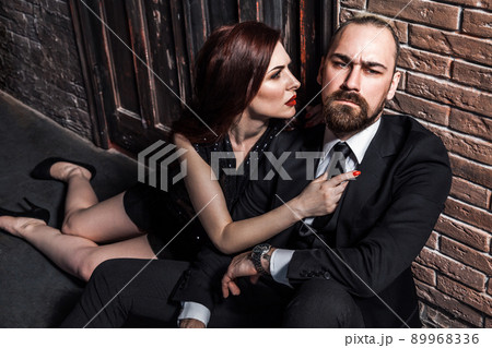 Man in suit and woman in black evening dress sitting on his lap. Foxy woman looks at him and wants to kiss. Studio shot, isolated on brick background 89968336